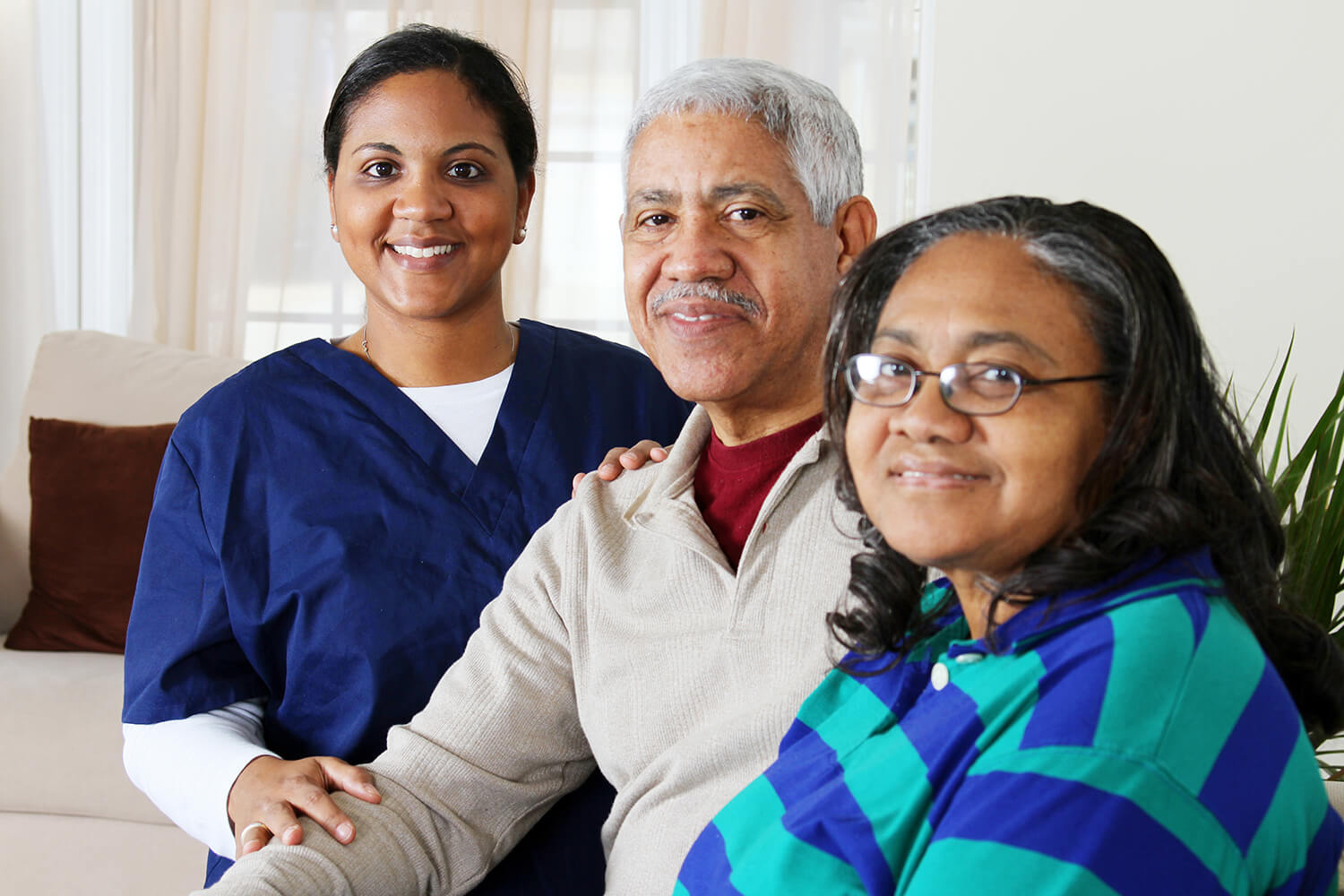 join-our-family Older man in grey sweater with maroon under shirt sits next to lady in blue and green stiped shirt with glasses while nurse in dark blue scrubs stands behind man's shoulder (all smiling)