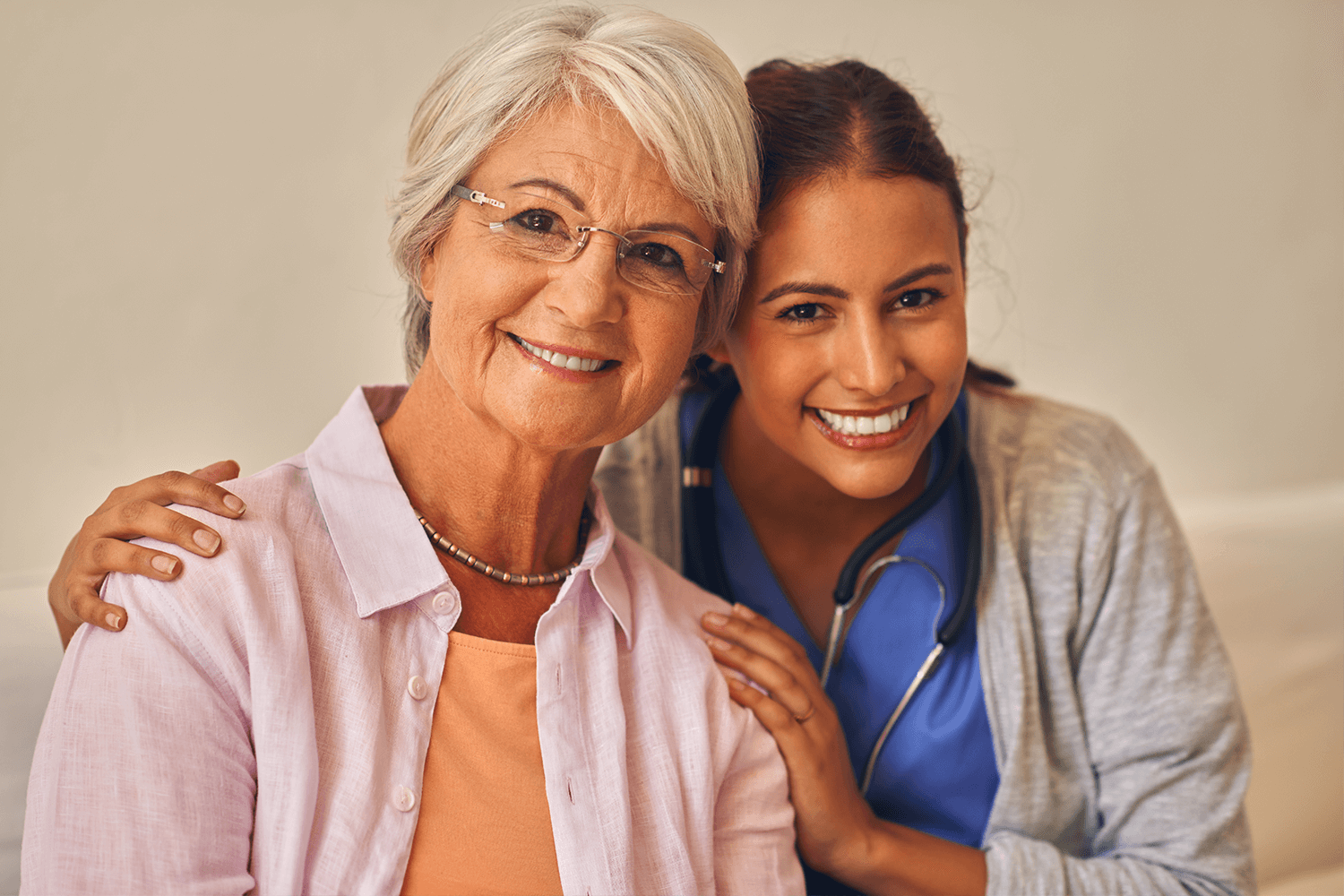 patient elderly lady wearing orange blouse with white rough silk over shirt, short grey hair parted over right eye with glasses and inquisitive smile leaning into a hug by a younger nurse with broad smile blue scrubs, stethiscope and grey cotton over shirt