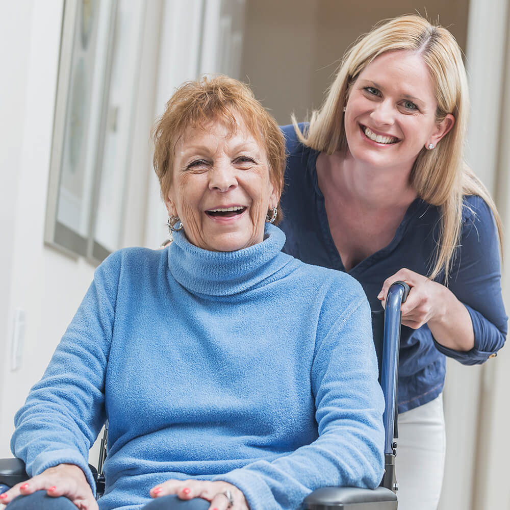 CDS Resources elderly lady in wheelchair wearing blue long sleeve turtleneck sweater with contentedly happy smile (possibly laughing) with younger lady leaning into the picture wearing darker blue V-neck shirt smiling for the picture.